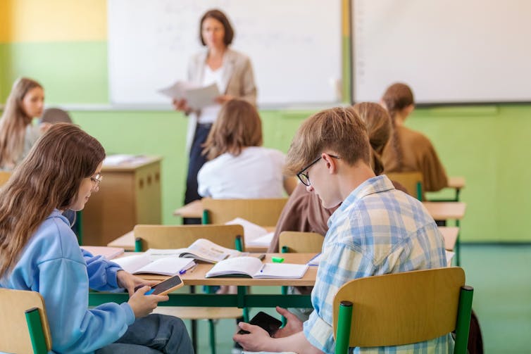 Students sit at a desk in a classroom using smartphones, while a teacher in the background looks on.
