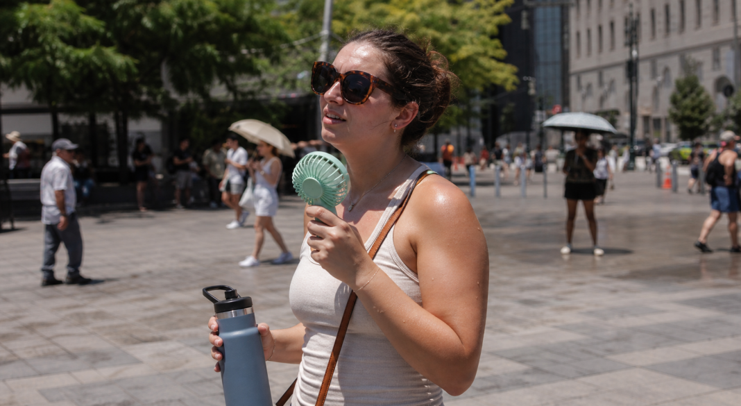 A woman using a handheld fan and carrying water during extreme summer heat in a city
